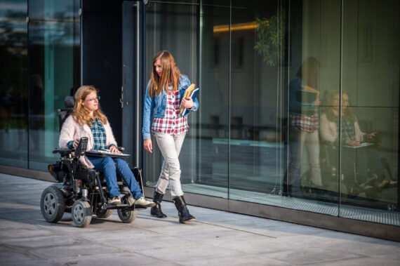 Disabled female student and her friend walking in campus, university building in background.