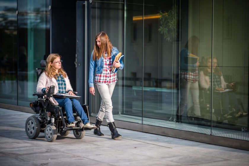 Disabled female student and her friend walking in campus, university building in background.