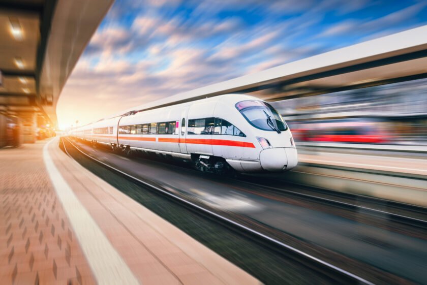 White modern speed train in motion on railway station at sunset. Train on railroad track with motion blur effect in Europe in evening. Railway platform. Industrial landscape. Railway tourism